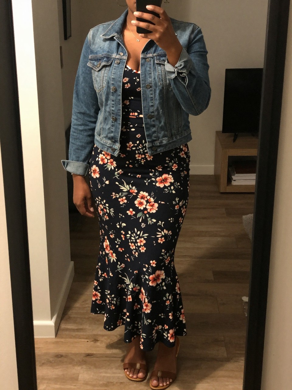 mirror selfie of a curvy black woman in a denim jacket and a floral printed mermaid skirt with flat sandals, casually holding her phone at her side, cropped at NECK so face doesn't show, in a city apartment, warm moody lighting, iPhone photo quality.