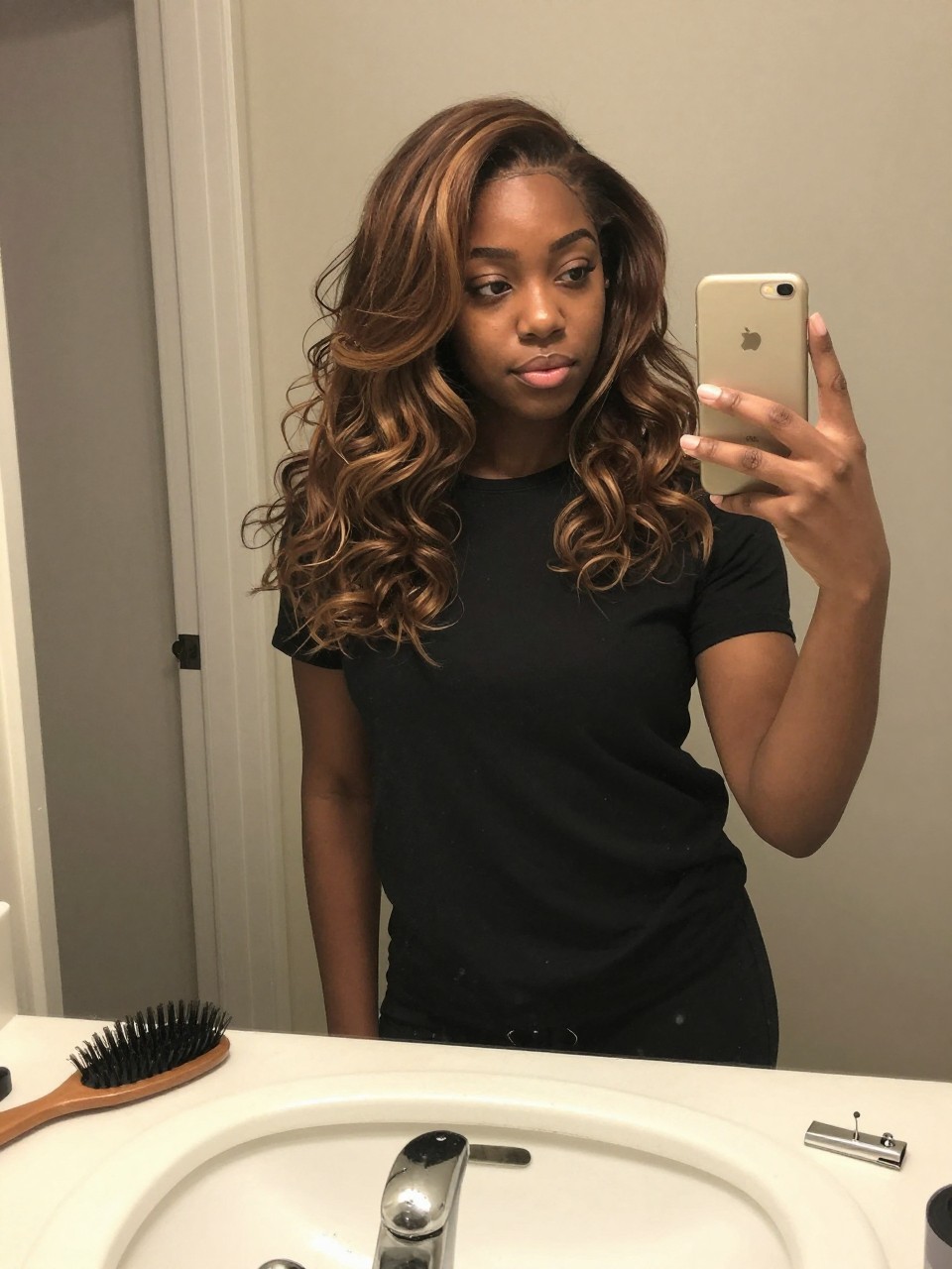 Photo of a Black woman wearing a wavy brown lob with mushroom brown highlights, candid mirror selfie, at a bathroom vanity mirror, soft indoor lighting, containing a small lived-in detail such as a hairbrush on the sink and a silver clip nearby, latest iPhone photo quality.