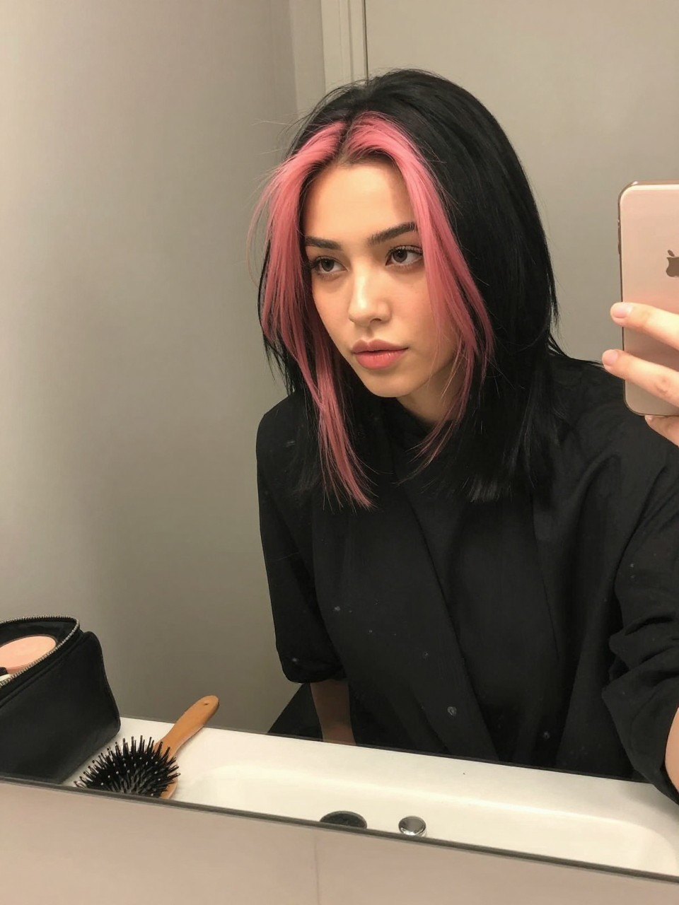 Photo of a woman wearing a sleek black lob with pink face-framing highlights, candid mirror selfie, at a bathroom vanity mirror, soft indoor lighting, containing a small lived-in detail such as a hairbrush on the vanity and a makeup bag nearby, latest iPhone photo quality.