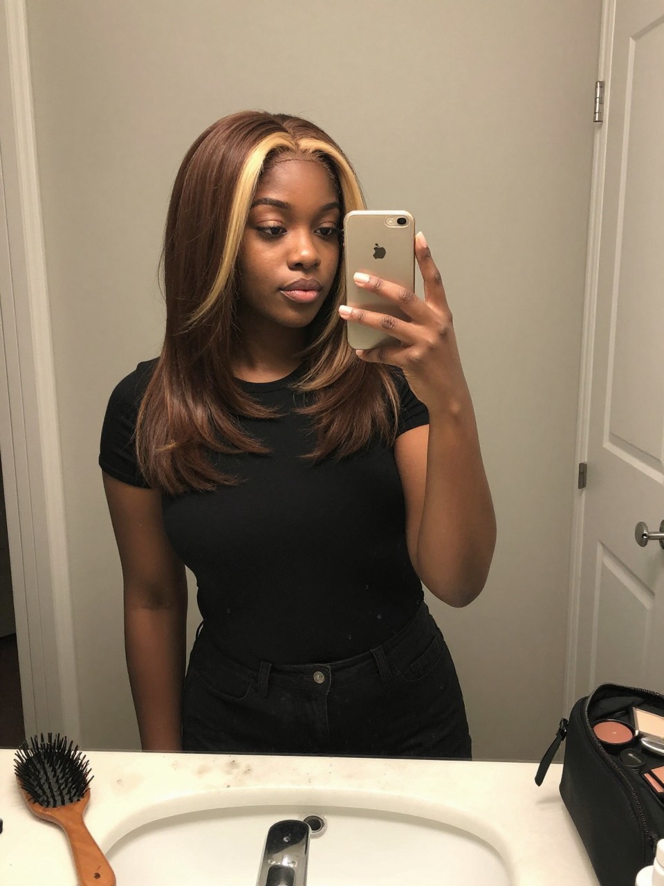 Photo of a black woman wearing a sleek espresso brunette lob with blonde face-framing highlights, candid mirror selfie, at a bathroom vanity mirror, soft indoor lighting, containing a small lived-in detail such as a hairbrush on the vanity and a makeup bag nearby, latest iPhone photo quality.