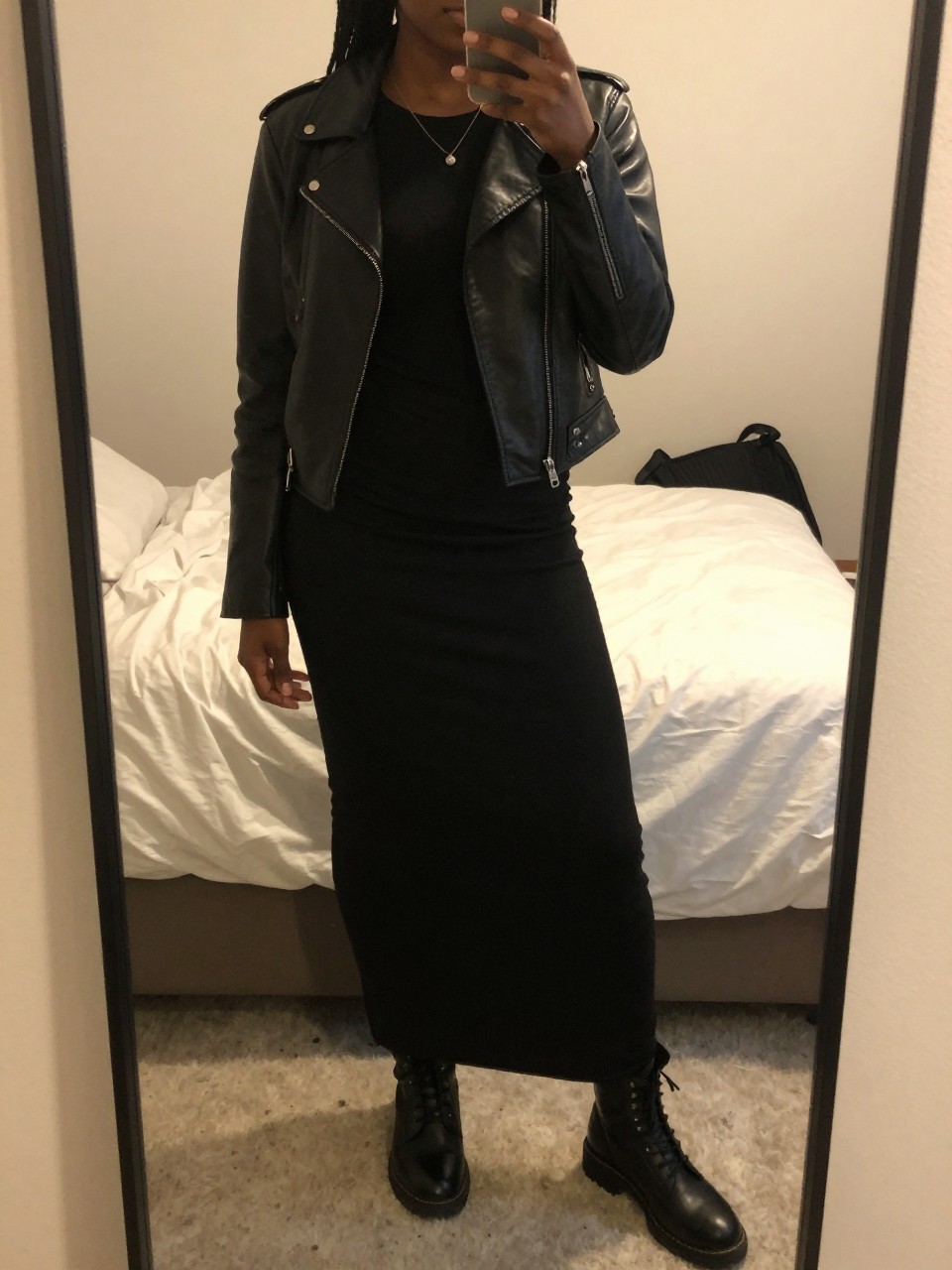 iPhone mirror selfie of a black woman in a black maxi skirt and leather jacket over a black tee with combat boots, standing with hand on hip, cropped at NECK so face doesn't show, in a feminine-themed bedroom, warm moody lighting, iPhone photo quality.
