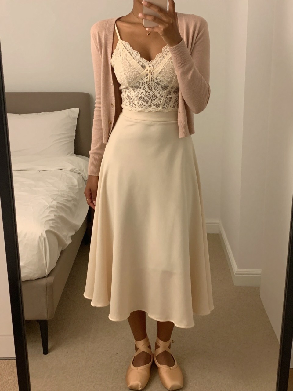 mirror selfie of a black woman wearing a cream lace cami under a soft blush cardigan tucked into a soft cream A-line midi skirt with nude ballet flats, one hand holding her phone at chest level, cropped at NECK so face doesn't show, in a feminine-themed bedroom, warm moody lighting, iPhone photo quality.