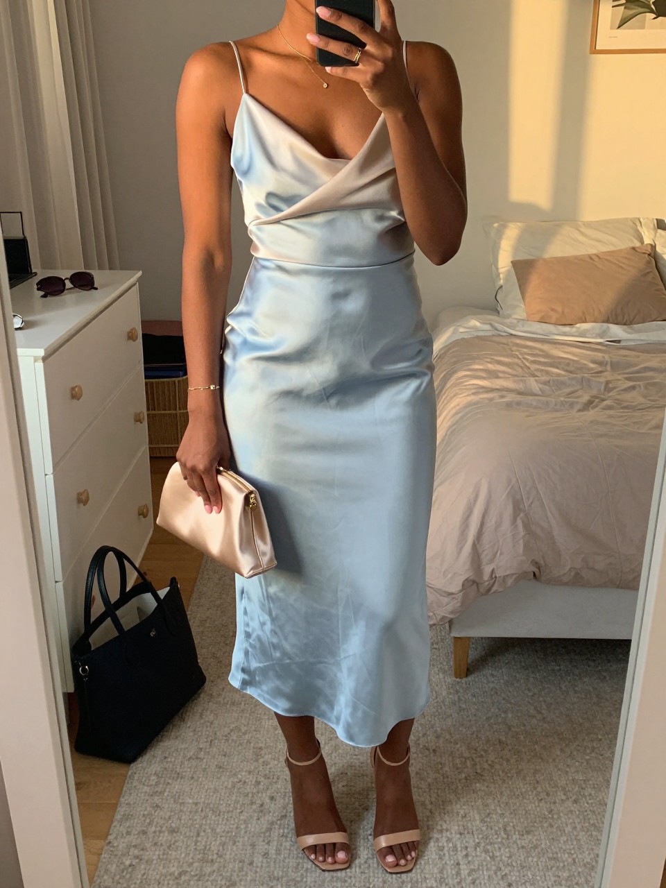 mirror selfie of a black woman wearing a light blue satin midi dress with a soft beige light wrap, nude leather heeled sandals, a delicate gold bracelet, and a champagne satin small clutch, holding a phone in front of a mirror, cropped at NECK so face doesn't show, with a natural casual selfie pose, in a feminine-themed bedroom, evening golden hour, including small lived-in details like a tote bag on the floor, sunglasses on a dresser, or soft bedding in the background, latest iPhone photo quality.