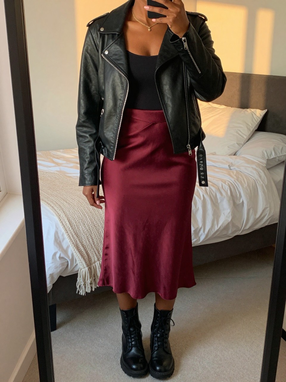 mirror selfie of a black woman wearing a black leather jacket over a black tank top with a burgundy satin slip midi skirt and black combat boots, one hand in her jacket pocket, cropped at NECK so face doesn't show, in a feminine-themed bedroom, evening golden hour, iPhone photo quality.