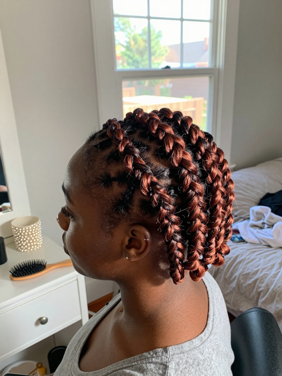 Photo of a Black woman wearing a flat twist crown with soft copper highlights woven through the twists, side profile view, in a sun-drenched bedroom, natural window light, containing a small lived-in detail such as pearl pins on the vanity and a hairbrush nearby, latest iPhone photo quality.