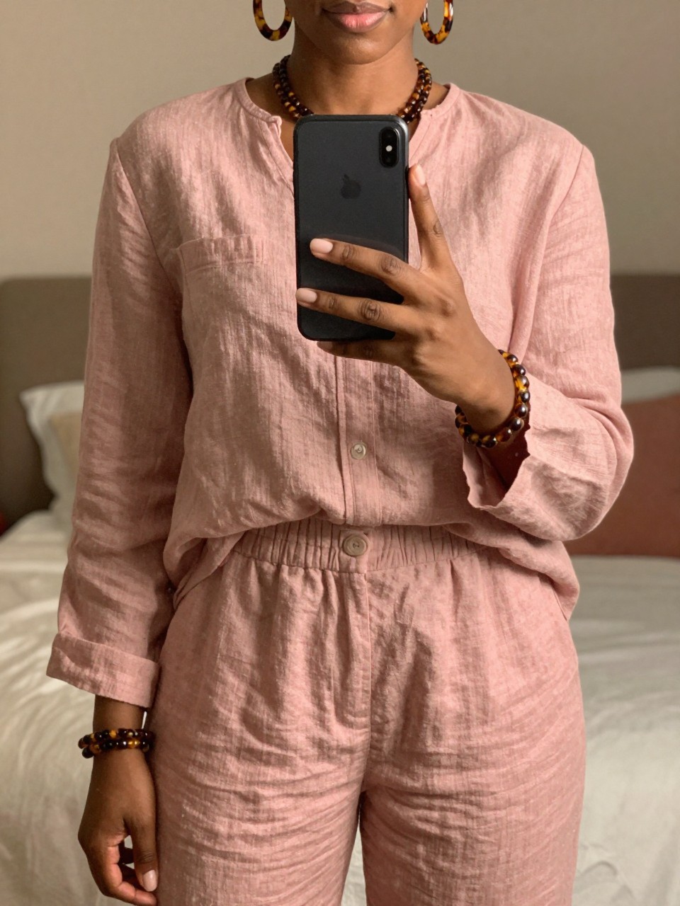 iPhone mirror selfie of a black woman in a dusty pink linen set with tortoiseshell accessories, holding phone at mid-chest, cropped at NECK so face doesn't show, in a feminine-themed bedroom, warm moody lighting, iPhone photo quality.