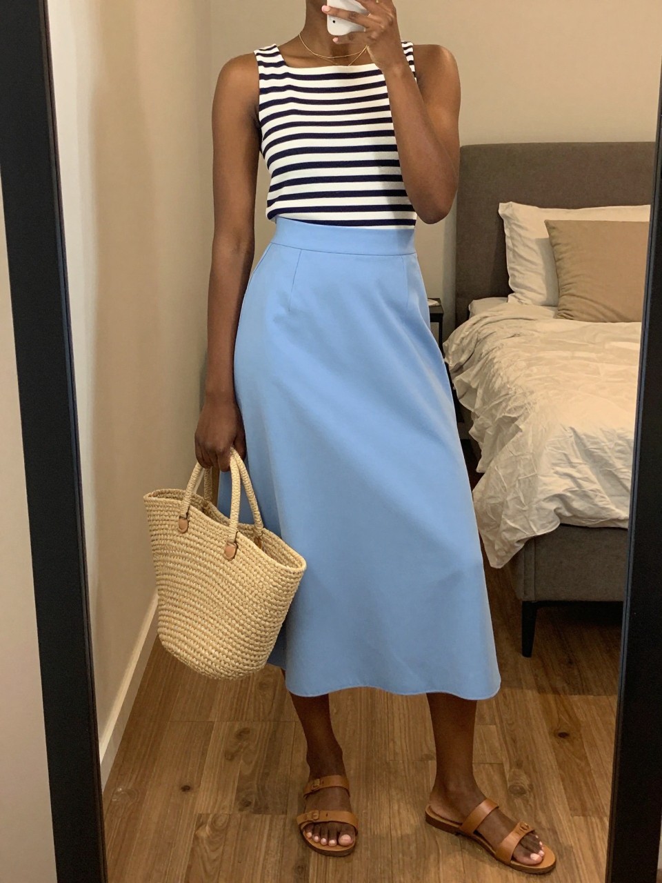 mirror selfie of a black woman wearing a navy and white striped boatneck top tucked into a sky blue A-line midi skirt with flat tan leather sandals and a straw market bag, thin gold necklace visible, standing in a relaxed elegant pose, cropped at NECK so face doesn't show, in a feminine-themed bedroom, warm moody lighting, iPhone photo quality.