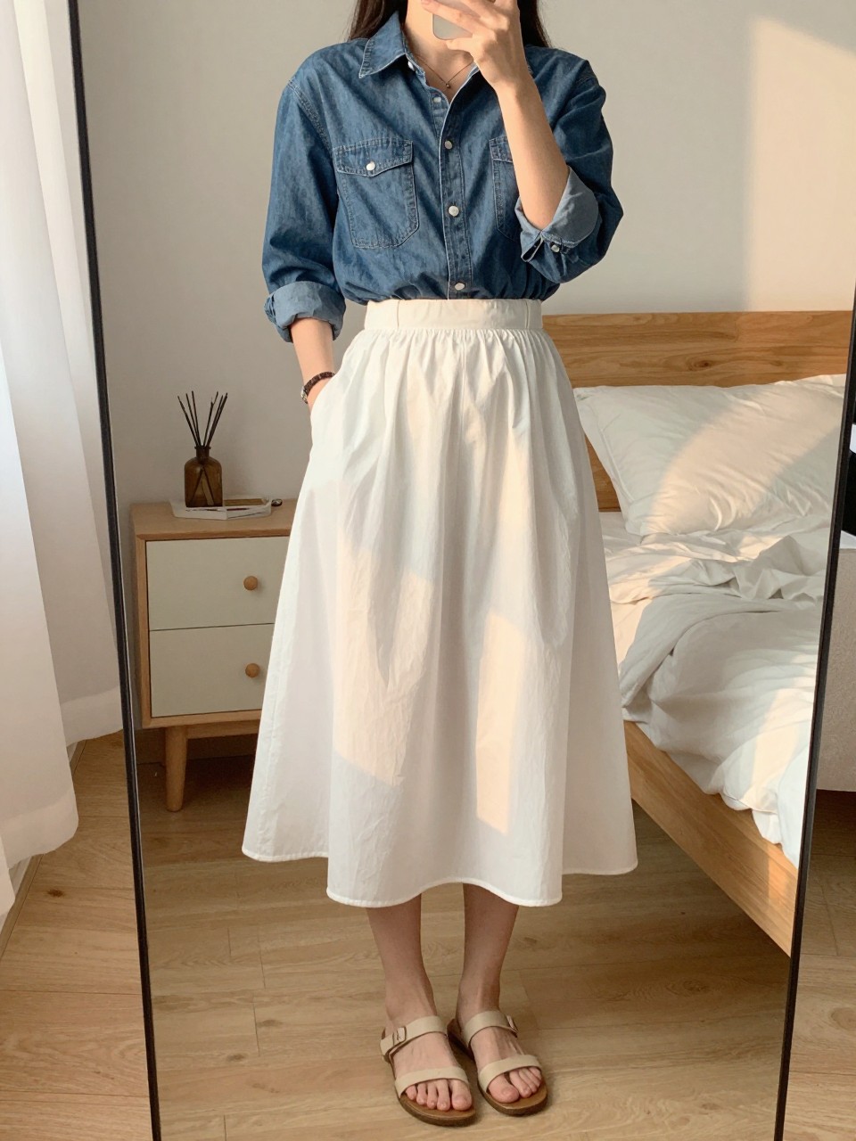 iPhone mirror selfie of a woman in a denim shirt and a white balloon skirt with flat sandals, standing with one hand in her pocket, cropped at NECK so face doesn't show, in a feminine-themed bedroom, evening golden hour, iPhone photo quality.
