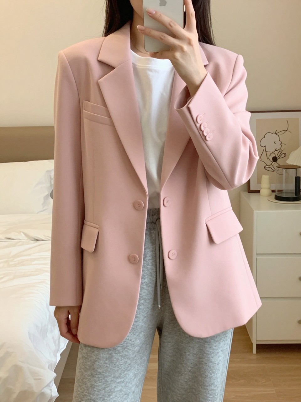 iPhone mirror selfie of a woman wearing a tailored pink blazer with a white tee and grey sweatpants, standing with phone in hand, cropped at NECK so face doesn't show, in a feminine-themed bedroom, warm moody lighting, iPhone photo quality.