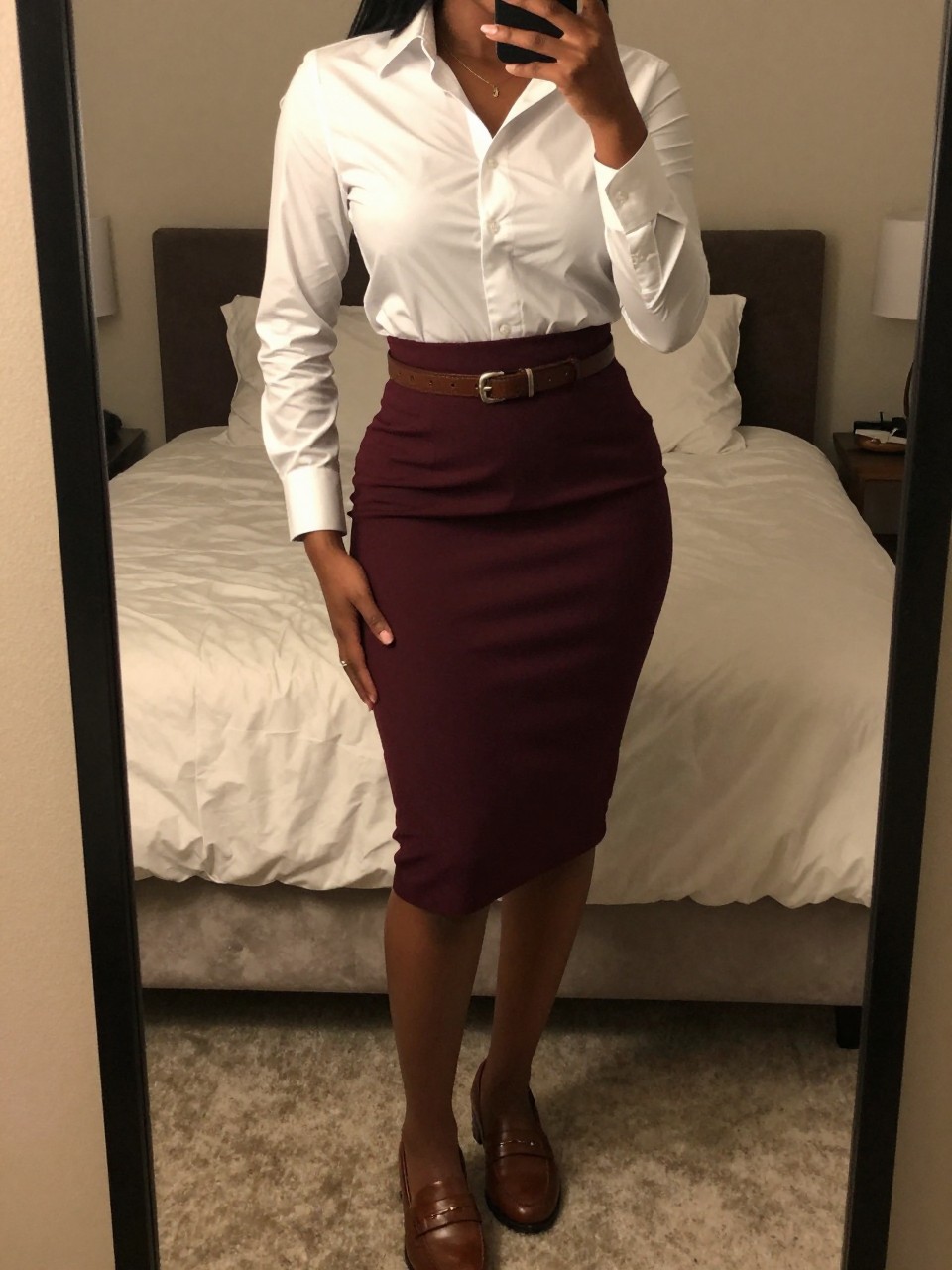 mirror selfie of a black woman wearing a white button-down shirt tucked into a deep burgundy pencil skirt with brown leather loafers and a matching belt, one hand in her pocket, cropped at NECK so face doesn't show, in a feminine-themed bedroom, warm moody lighting, iPhone photo quality.