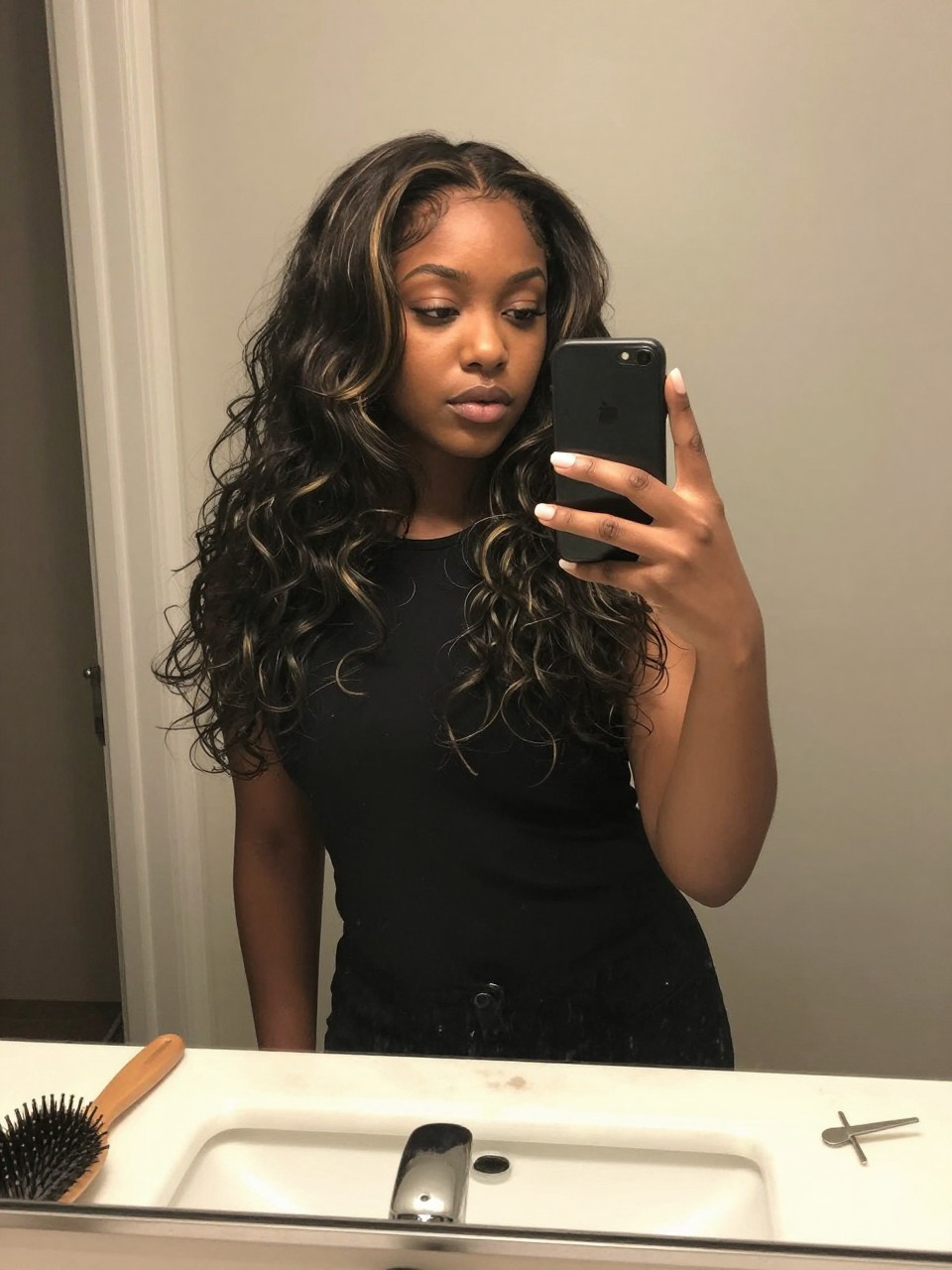 Photo of a black woman wearing a wavy black lob with ash blonde highlights, candid mirror selfie, at a bathroom vanity mirror, soft indoor lighting, containing a small lived-in detail such as a hairbrush on the sink and a silver clip nearby, latest iPhone photo quality.