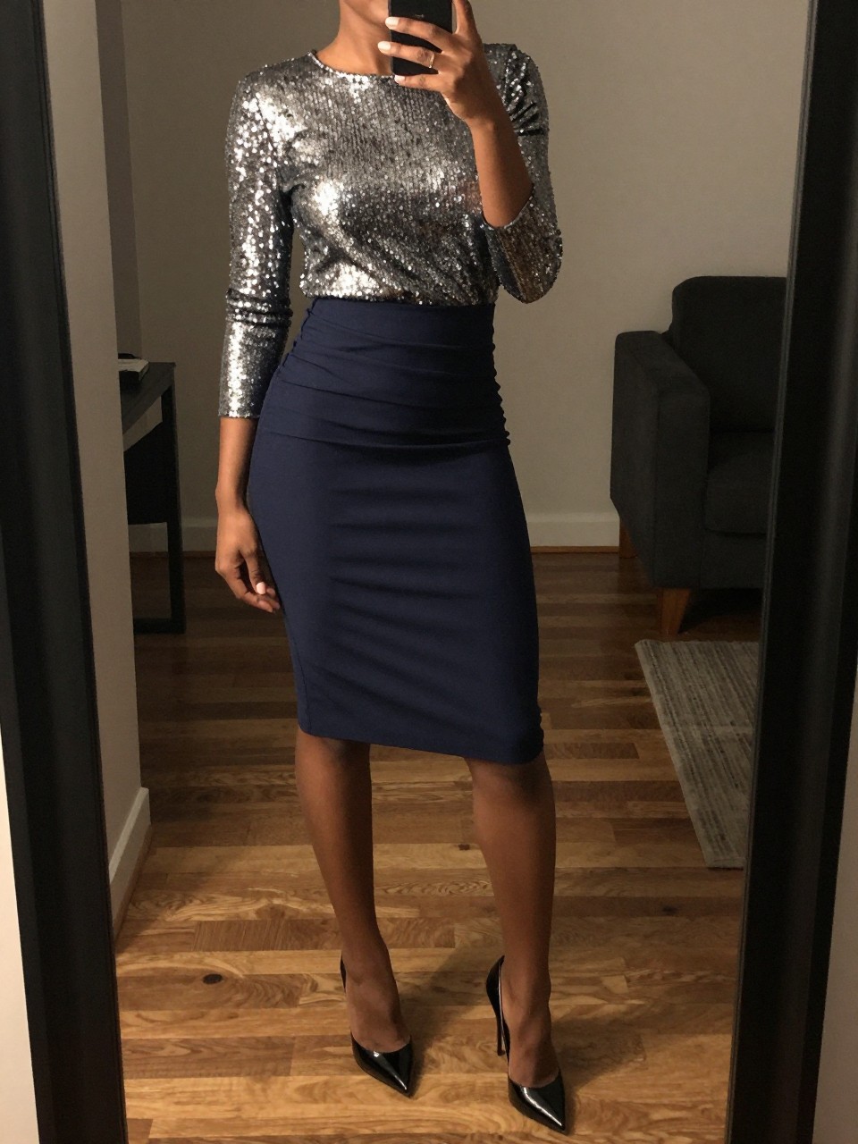 iPhone mirror selfie of a black woman wearing a silver sequin top tucked into a navy crepe pencil skirt with black patent stiletto heels, one hand holding her phone at her side, cropped at NECK so face doesn't show, in a city apartment, warm moody lighting, iPhone photo quality.