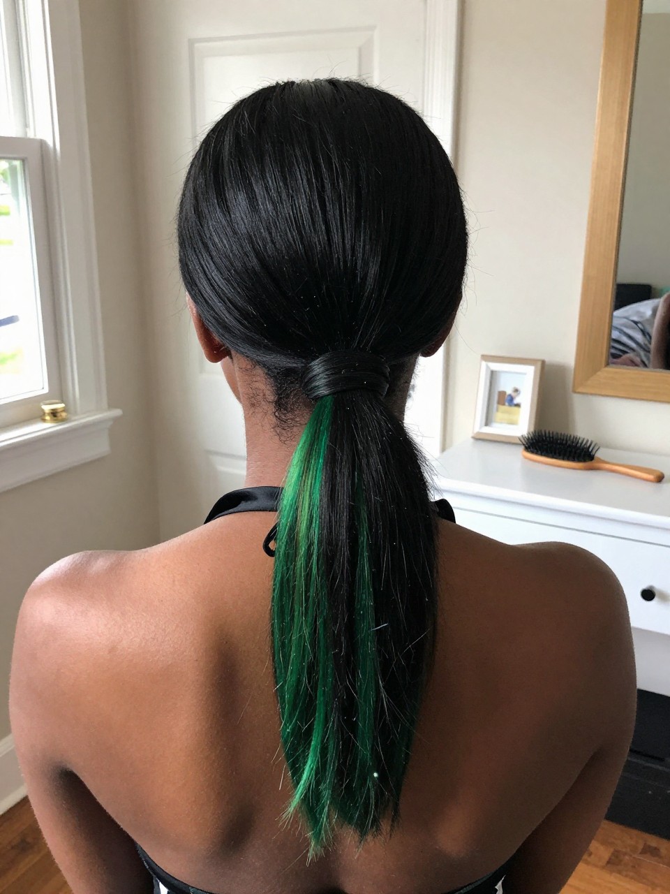 Photo of a black woman wearing a sleek low ponytail with black hair and green highlights through the tail and ends, back-of-head view to highlight details, in a sun-drenched bedroom, natural window light, containing a small lived-in detail such as a black ribbon tied at the base and a hairbrush on the vanity, latest iPhone photo quality.