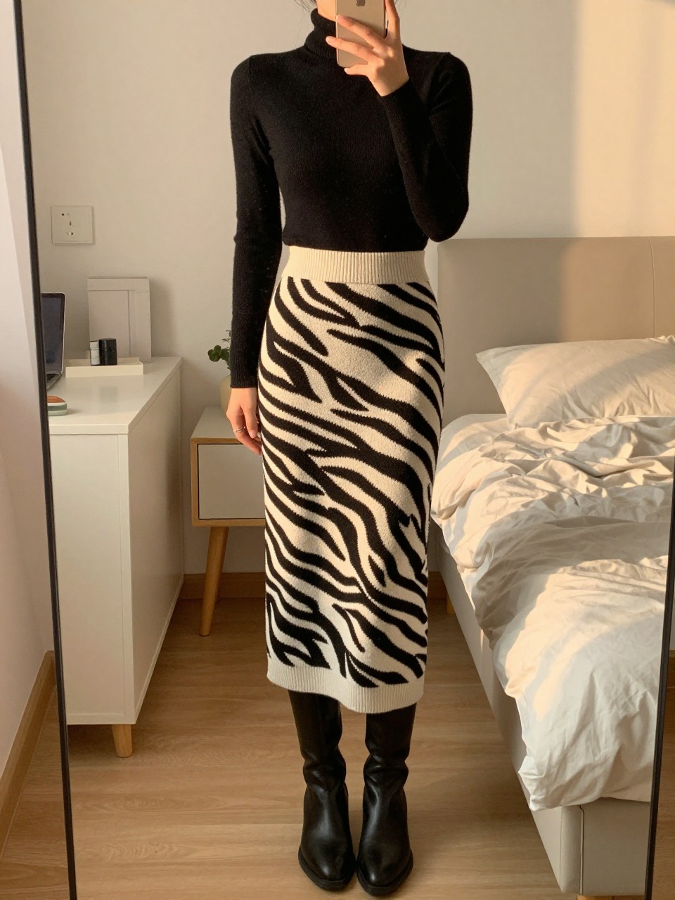 mirror selfie of a woman in a black turtleneck and a zebra print knit long skirt with knee-high boots, standing with phone in hand, cropped at NECK so face doesn't show, in a feminine-themed bedroom, evening golden hour, iPhone photo quality.