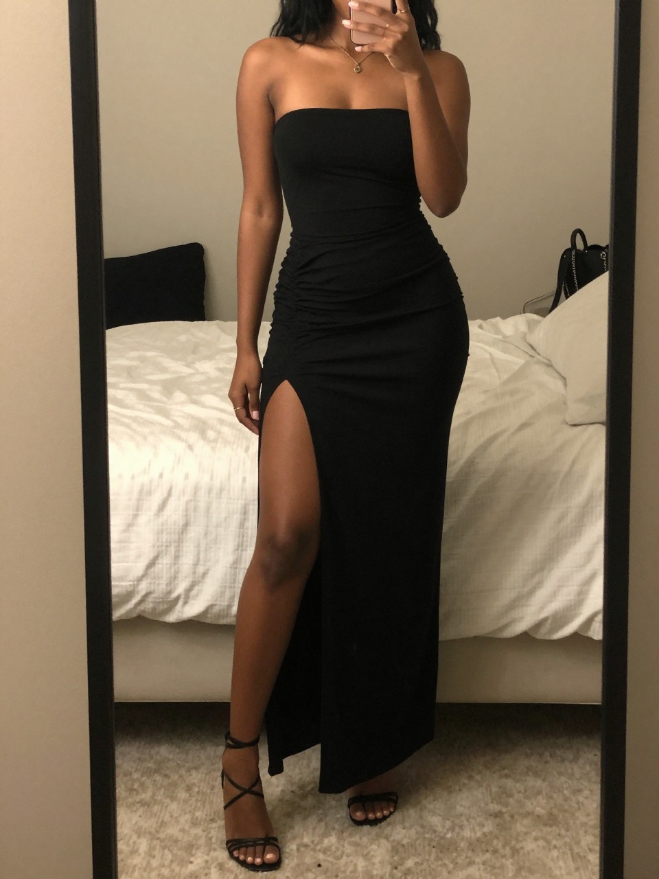 iPhone mirror selfie of a black woman in a black maxi skirt with a slit and strappy heels, standing with leg slightly exposed through slit, cropped at NECK so face doesn't show, in a feminine-themed bedroom, warm moody lighting, iPhone photo quality.