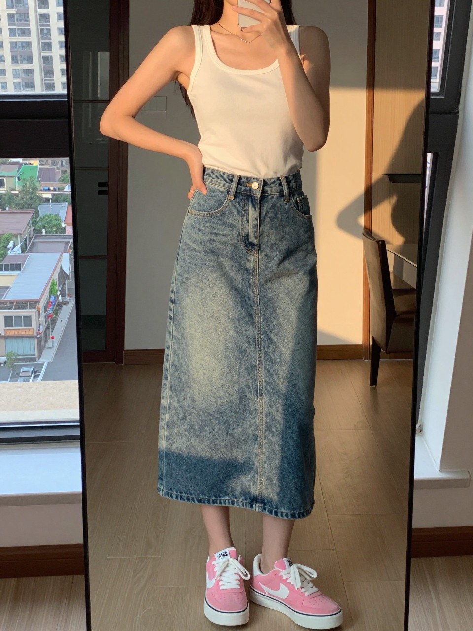 iPhone mirror selfie of a woman in a denim skirt and white tank with pink sneakers, standing with hand on hip, cropped at NECK so face doesn't show, in a city apartment, evening golden hour, iPhone photo quality.