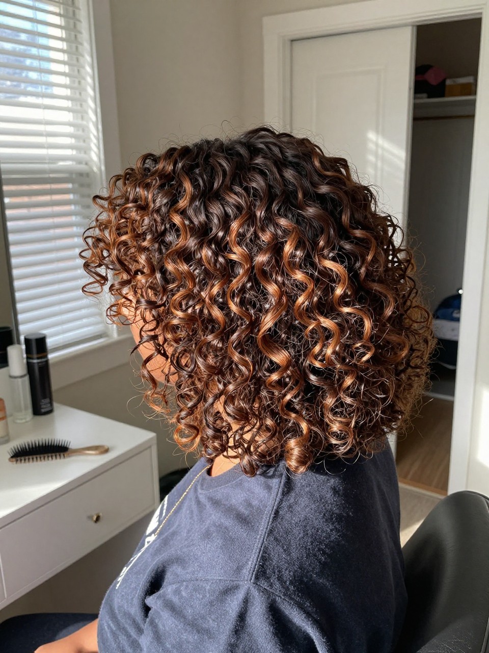 Photo of a black woman wearing curly brunette hair with cinnamon brown highlights and lifted roots, back-of-head view to show the warm highlighted curls, sun-drenched bedroom setting, natural window light, with a hair pick on the vanity and a slightly open closet behind, latest iPhone photo quality.