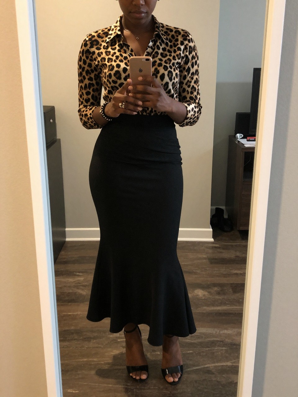 mirror selfie of a black woman in a leopard print blouse and a black mermaid skirt with kitten heels, holding her phone at waist level, cropped at NECK so face doesn't show, in a city apartment, warm moody lighting, iPhone photo quality.