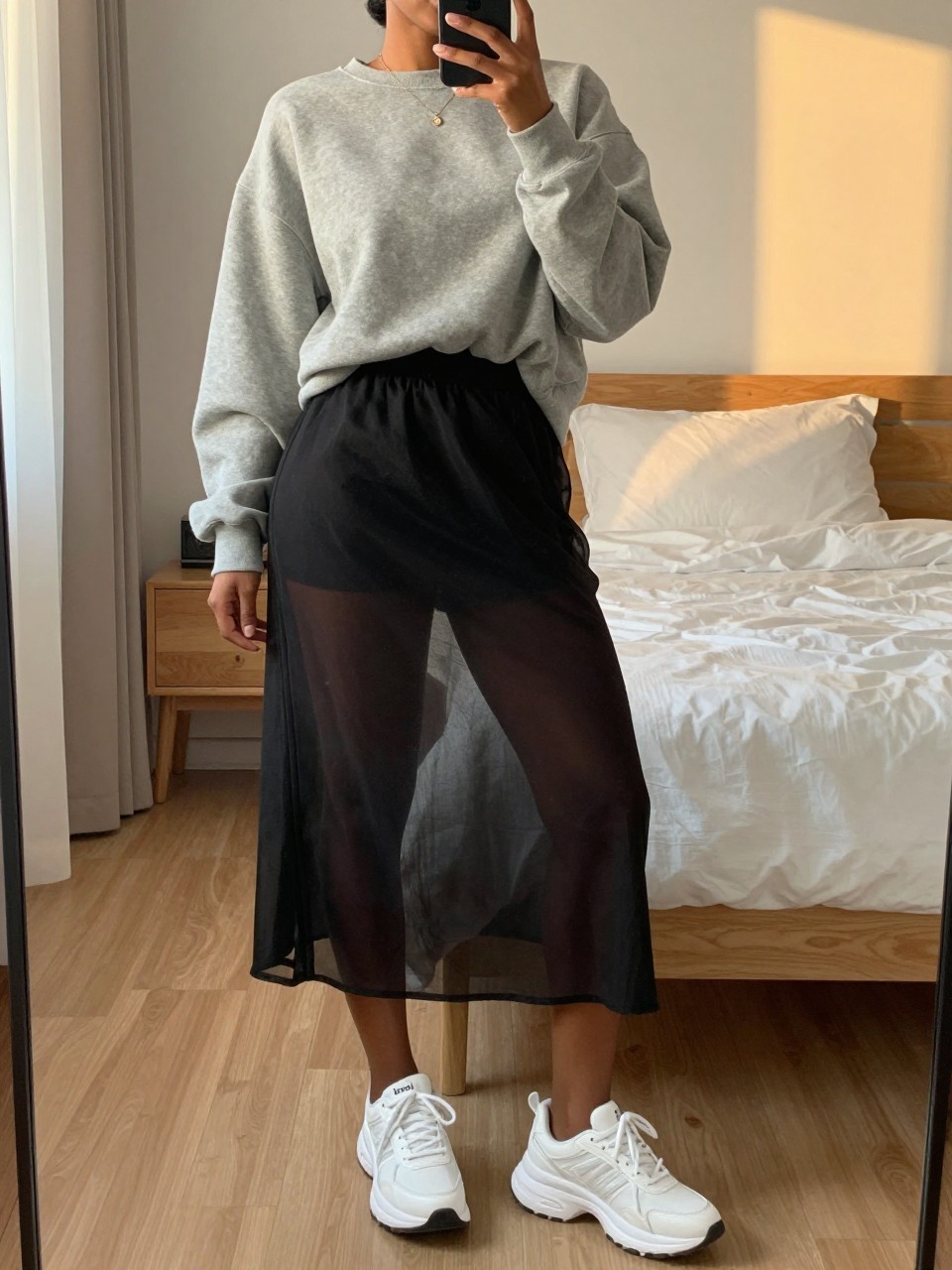 mirror selfie of a black woman wearing an oversized gray sweatshirt half-tucked into a black sheer mesh midi skirt with chunky white sneakers, one hand holding her phone at her side, cropped at NECK so face doesn't show, in a feminine-themed bedroom, evening golden hour, iPhone photo quality.
