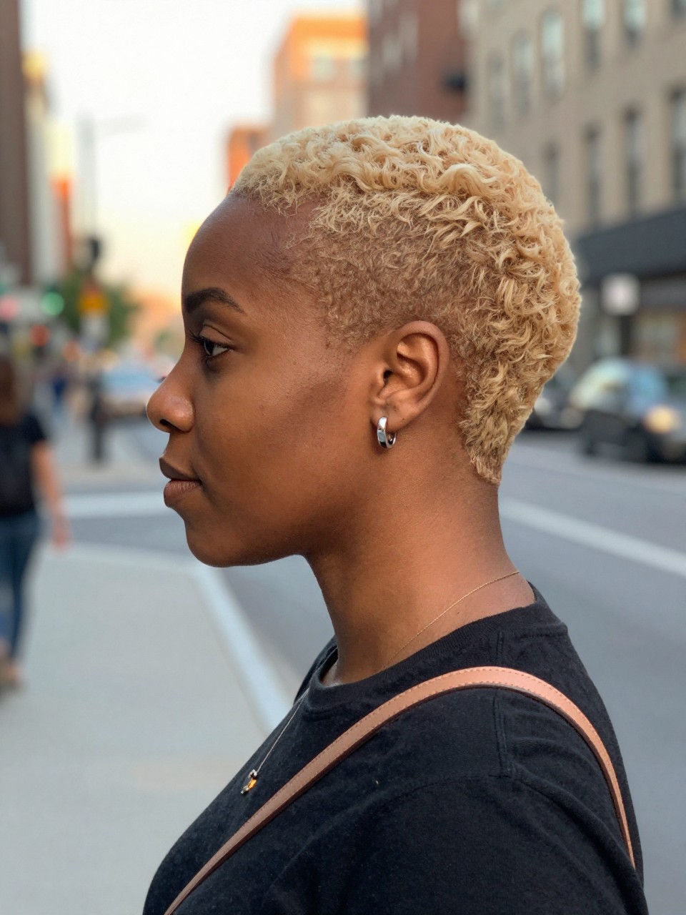 Photo of a black woman wearing a blunt ginger hairstyle with a soft highlight melt, side profile view, in a softly blurred city street background, golden hour glow, containing a small lived-in detail such as statement earrings peeking through and a crossbody strap on the shoulder, latest iPhone photo quality.