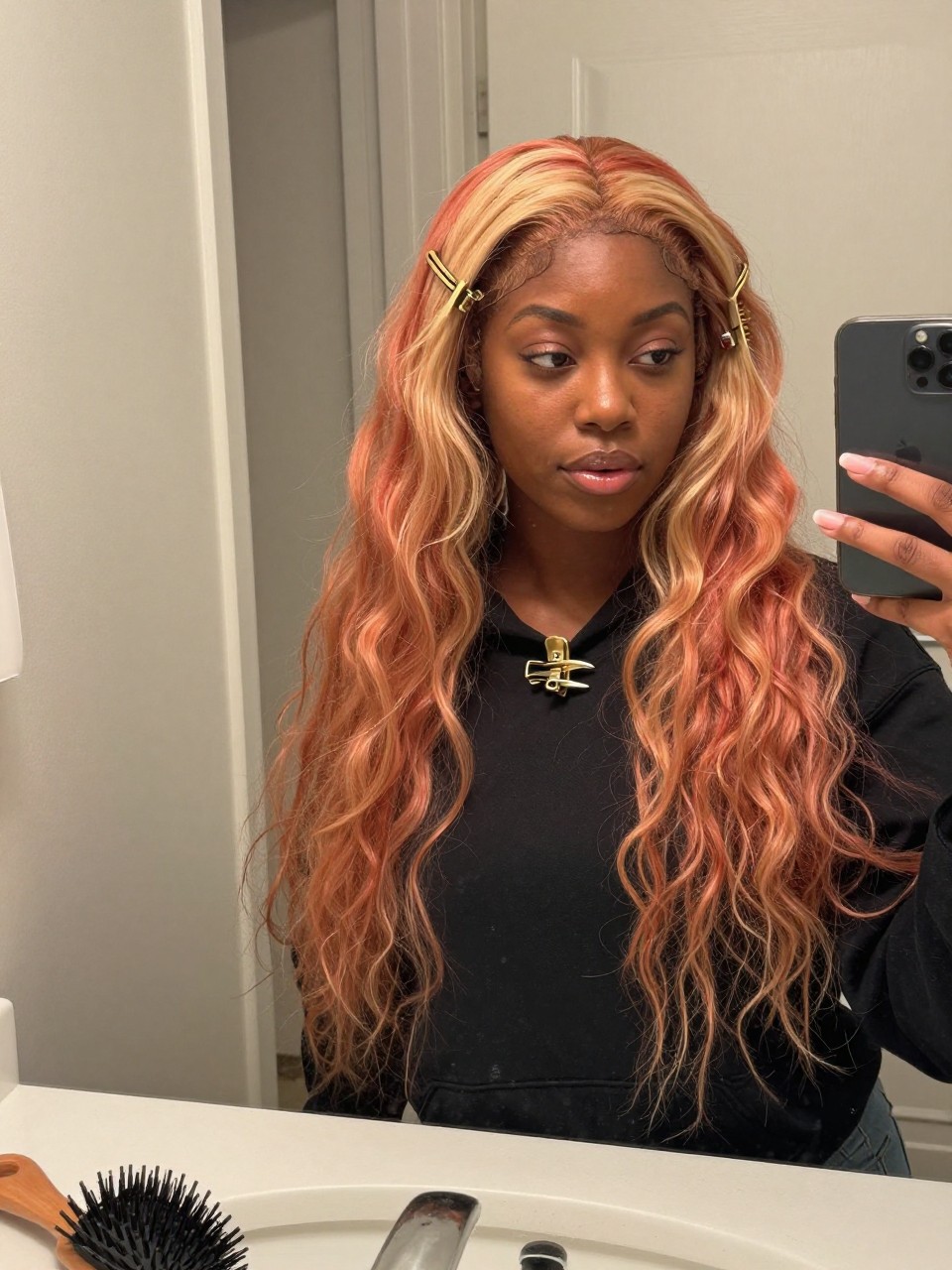 Photo of a black woman wearing a loose claw clip twist with strawberry blonde hair and warm blonde highlight pieces falling softly, candid mirror selfie, at a bathroom vanity mirror, soft indoor lighting, containing a small lived-in detail such as a claw clip clipped to the collar before use and a hairbrush on the sink, latest iPhone photo quality.