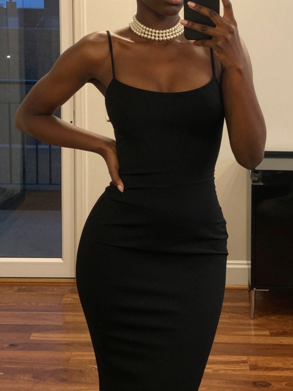 mirror selfie of a black woman in a sheath black dress and pearl choker, hand on hip, cropped at NECK so face doesn't show, in a city apartment, warm moody lighting, iPhone photo quality.