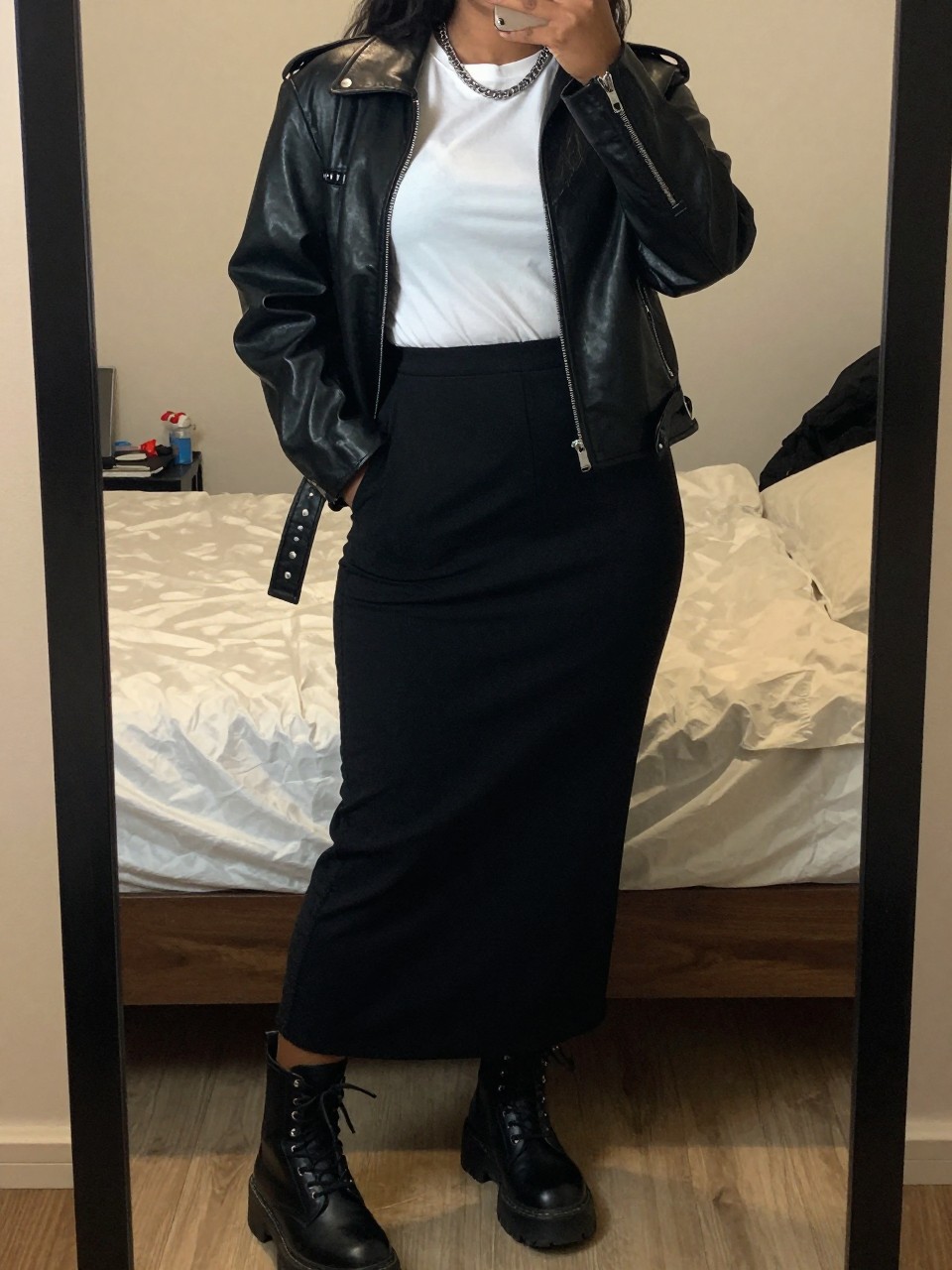 mirror selfie of a black curvy woman wearing a black leather jacket over a white tee tucked into a black cotton maxi skirt with black chunky combat boots and a silver chain necklace, standing in an edgy pose with hands in pockets, cropped at NECK so face doesn't show, in a feminine-themed bedroom, warm moody lighting, iPhone photo quality.