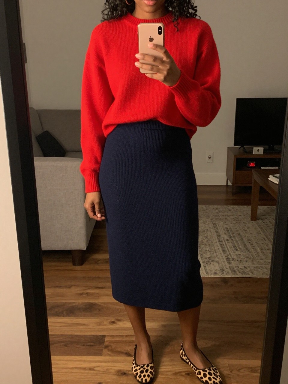 iPhone mirror selfie of a black woman wearing a red cashmere sweater tucked into a navy knit midi skirt with leopard print flats, one hand holding her phone at chest level, cropped at NECK so face doesn't show, in a city apartment, warm moody lighting, iPhone photo quality.