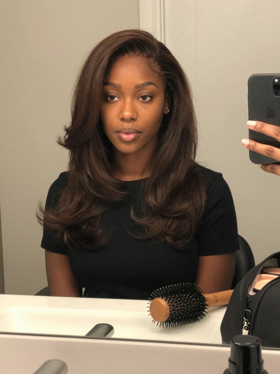 Photo of a black woman wearing deep chocolate brown hair with soft face-framing layers in a smooth blowout, candid mirror selfie, bathroom vanity mirror setting, soft indoor lighting, with a blowout brush on the counter and a makeup bag nearby, latest iPhone photo quality.