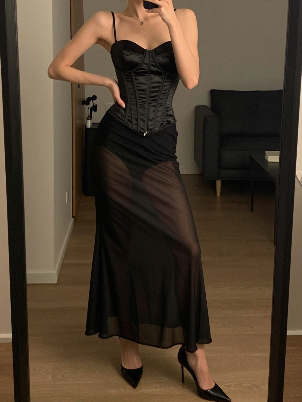 mirror selfie of a woman wearing a black satin corset top with a black sheer mesh maxi skirt and black stiletto heels, one hand on her hip, cropped at NECK so face doesn't show, in a city apartment, warm moody lighting, iPhone photo quality.