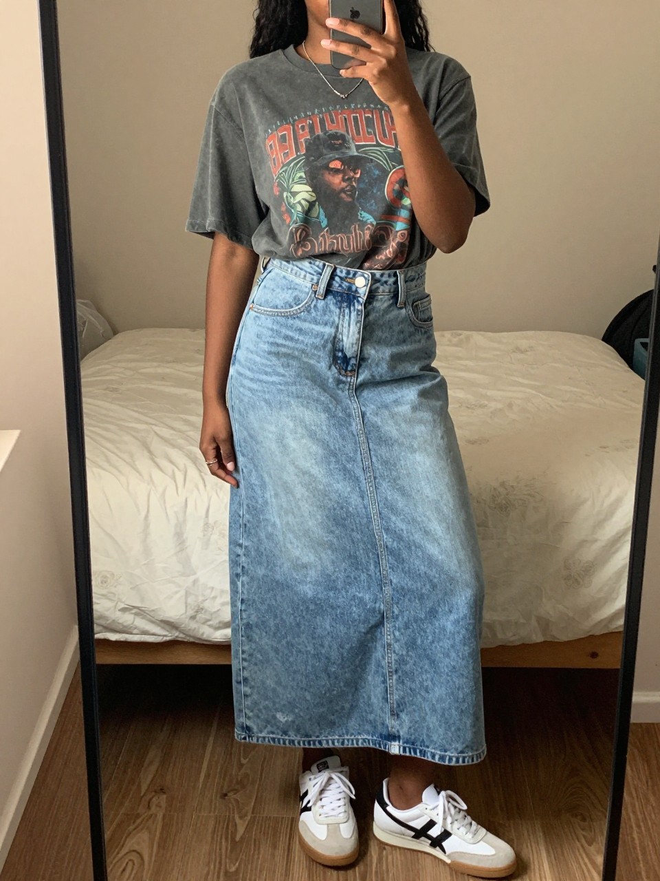 iPhone mirror selfie of a black woman wearing a faded vintage band tee tucked loosely into a light-wash denim maxi skirt with retro white sneakers and a baseball cap, standing in a casual stance, cropped at NECK so face doesn't show, in a feminine-themed bedroom, warm moody lighting, iPhone photo quality.