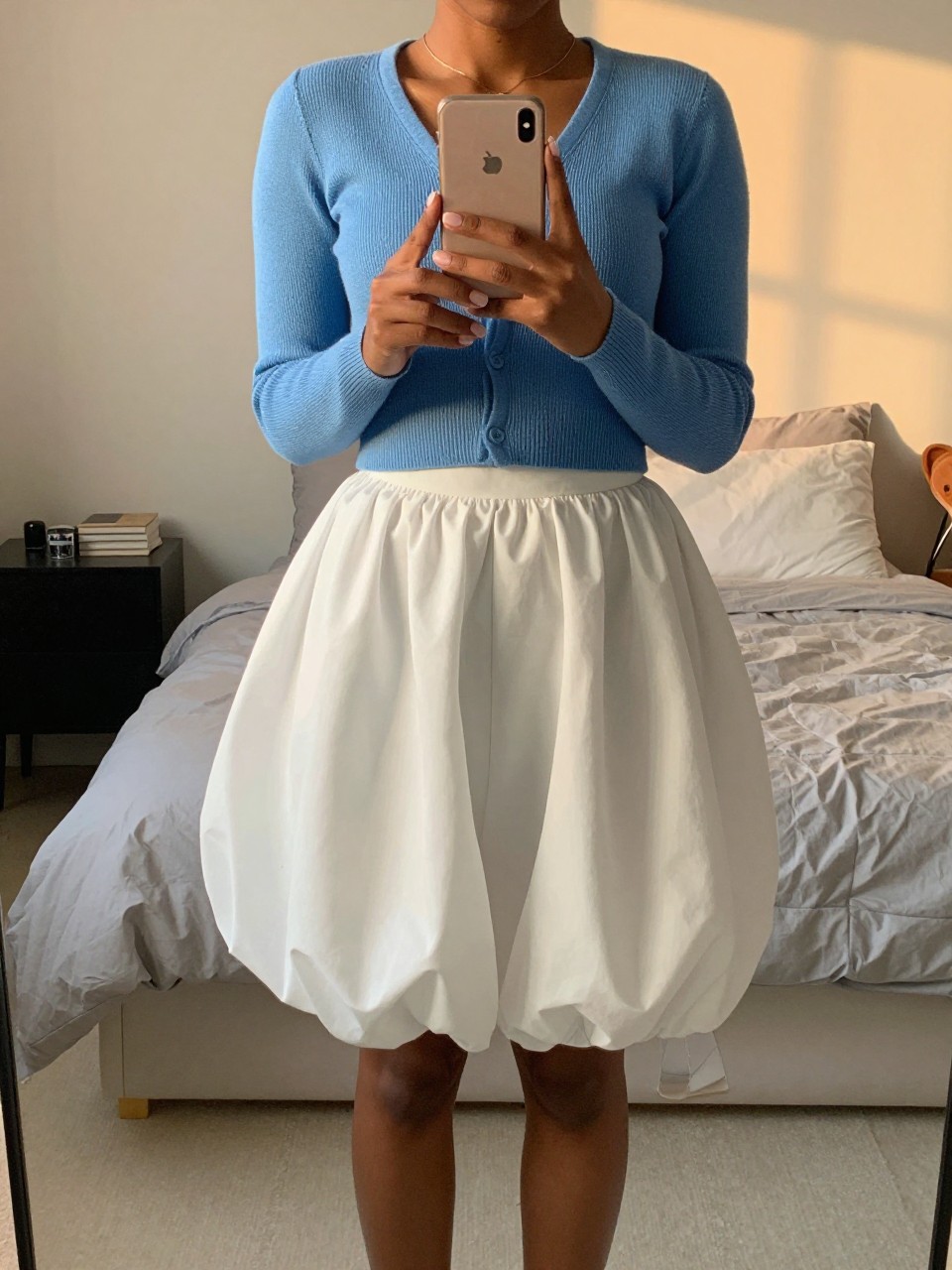mirror selfie of a black woman in a blue cropped cardigan and a white balloon skirt with Mary Janes, gently holding her phone at waist level, cropped at NECK so face doesn't show, in a feminine-themed bedroom, evening golden hour, iPhone photo quality.