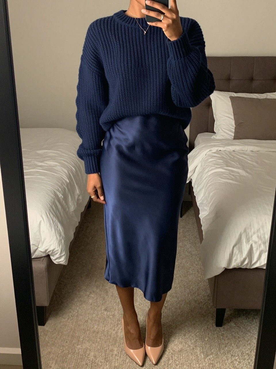mirror selfie of a black woman wearing a navy blue chunky knit sweater tucked into a matching navy blue satin midi skirt creating a monochrome look, with nude pointed heels and minimal gold jewelry, standing in a sophisticated pose, cropped at NECK so face doesn't show, in a feminine-themed bedroom, warm moody lighting, iPhone photo quality.