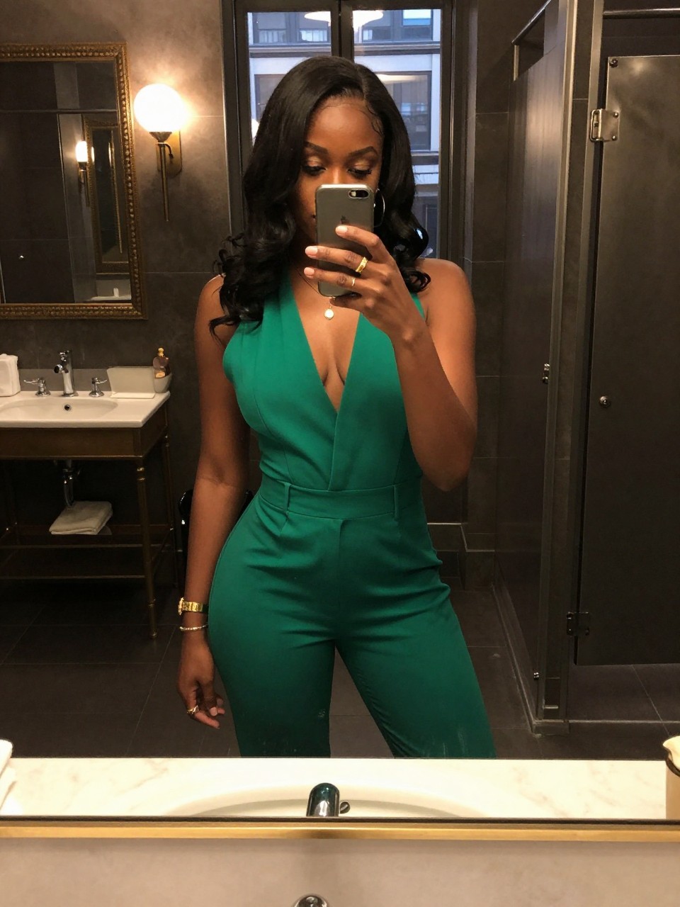 mirror selfie of a black woman in an emerald green jumpsuit with gold accessories, standing in a chic restaurant bathroom, cropped at NECK so face doesn't show, in a city apartment, warm moody lighting, iPhone photo quality.