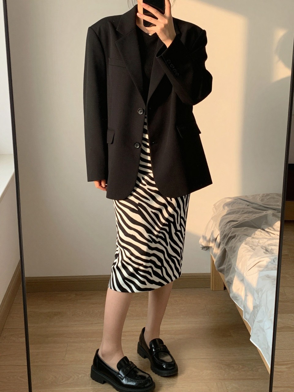 mirror selfie of a woman in an oversized black blazer and a zebra print pencil skirt with loafers, standing with her phone in hand, cropped at NECK so face doesn't show, in a feminine-themed bedroom, evening golden hour, iPhone photo quality.