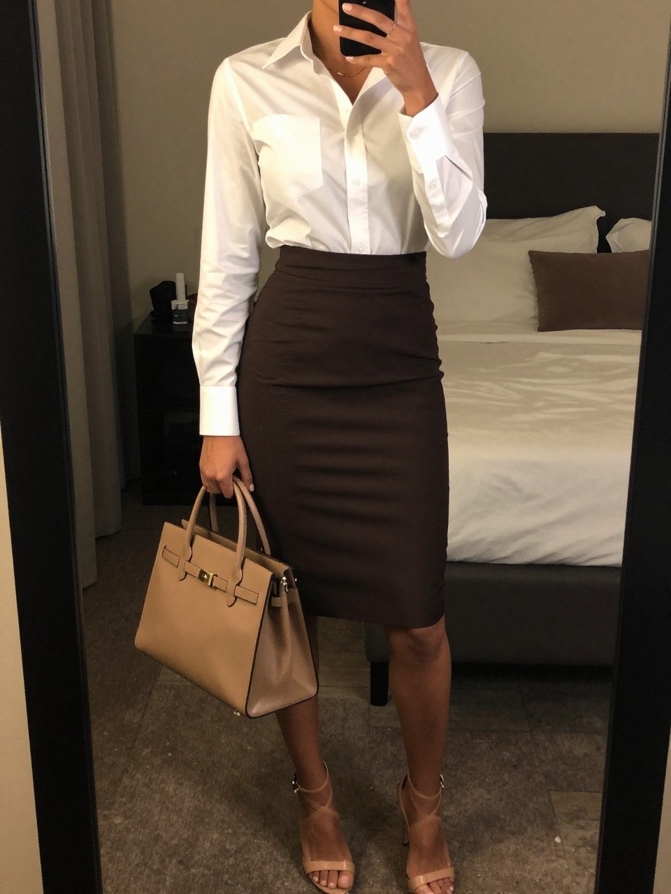 mirror selfie of a bkack woman wearing a crisp white button-down shirt tucked into a dark chocolate brown pencil skirt with nude strappy heeled sandals and a structured tan top-handle bag, standing in a professional pose, cropped at NECK so face doesn't show, in a feminine-themed bedroom, warm moody lighting, iPhone photo quality.