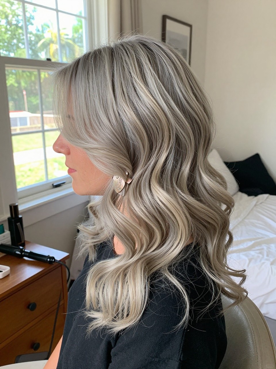 Photo of a woman wearing side part grey blonde waves with frosted ashy ends, side profile view, in a sun-drenched bedroom, natural window light, containing a small lived-in detail such as a decorative clip on one side and a curling iron resting on the nightstand, latest iPhone photo quality.
