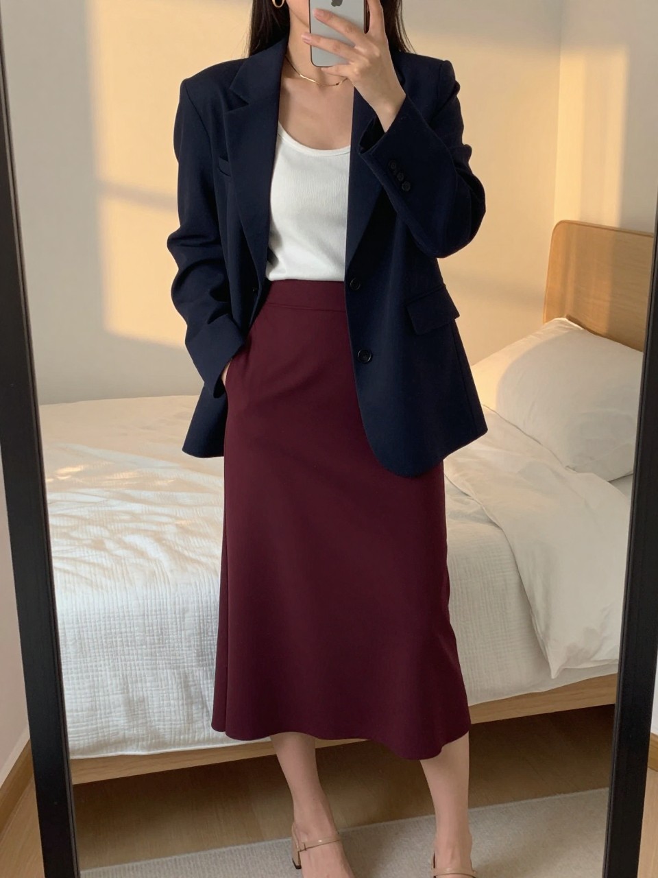 iPhone mirror selfie of a woman wearing a navy blazer over a white tank top tucked into a deep burgundy midi skirt with gold hoop earrings and nude block-heel sandals, one hand in her blazer pocket, cropped at NECK so face doesn't show, in a feminine-themed bedroom, evening golden hour, iPhone photo quality.