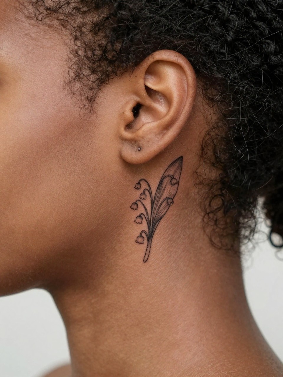 Image Prompt: "Extreme close-up of a small lily of the valley tattoo behind the ear of black feminine woman, fine line black ink floral design with tiny hanging flowers and soft shading, realistic skin texture, a few loose hair strands framing the placement, minimal feminine styling, tight crop, clean soft background, tattoo is the hero of the image (no watermarks on images)"