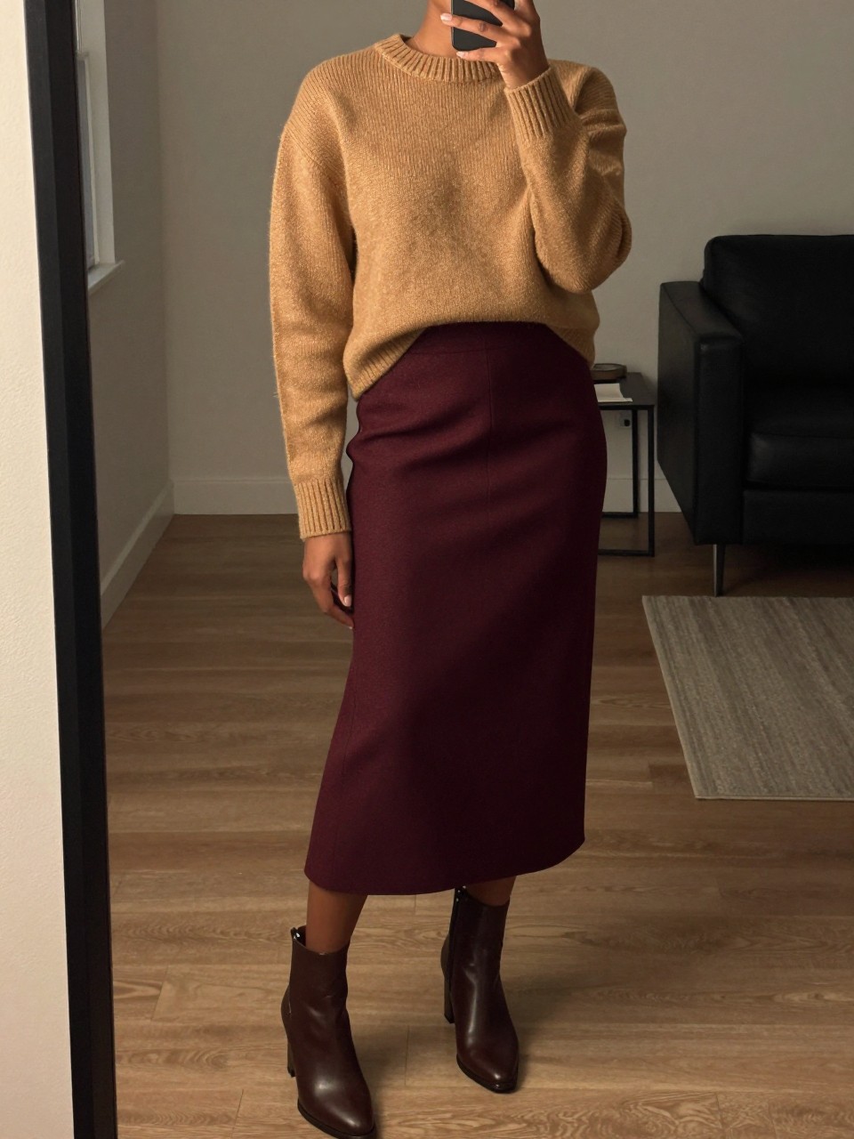 mirror selfie of a black woman wearing a camel crewneck sweater tucked into a deep burgundy wool midi skirt with brown block-heel ankle boots, one hand adjusting her sleeve, cropped at NECK so face doesn't show, in a city apartment, warm moody lighting, iPhone photo quality.