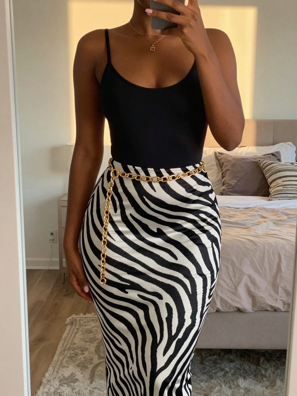 mirror selfie of a black woman in a black bodysuit and a zebra print midi skirt with a gold chain belt, hand resting on the chain, cropped at NECK so face doesn't show, in a feminine-themed bedroom, evening golden hour, iPhone photo quality.