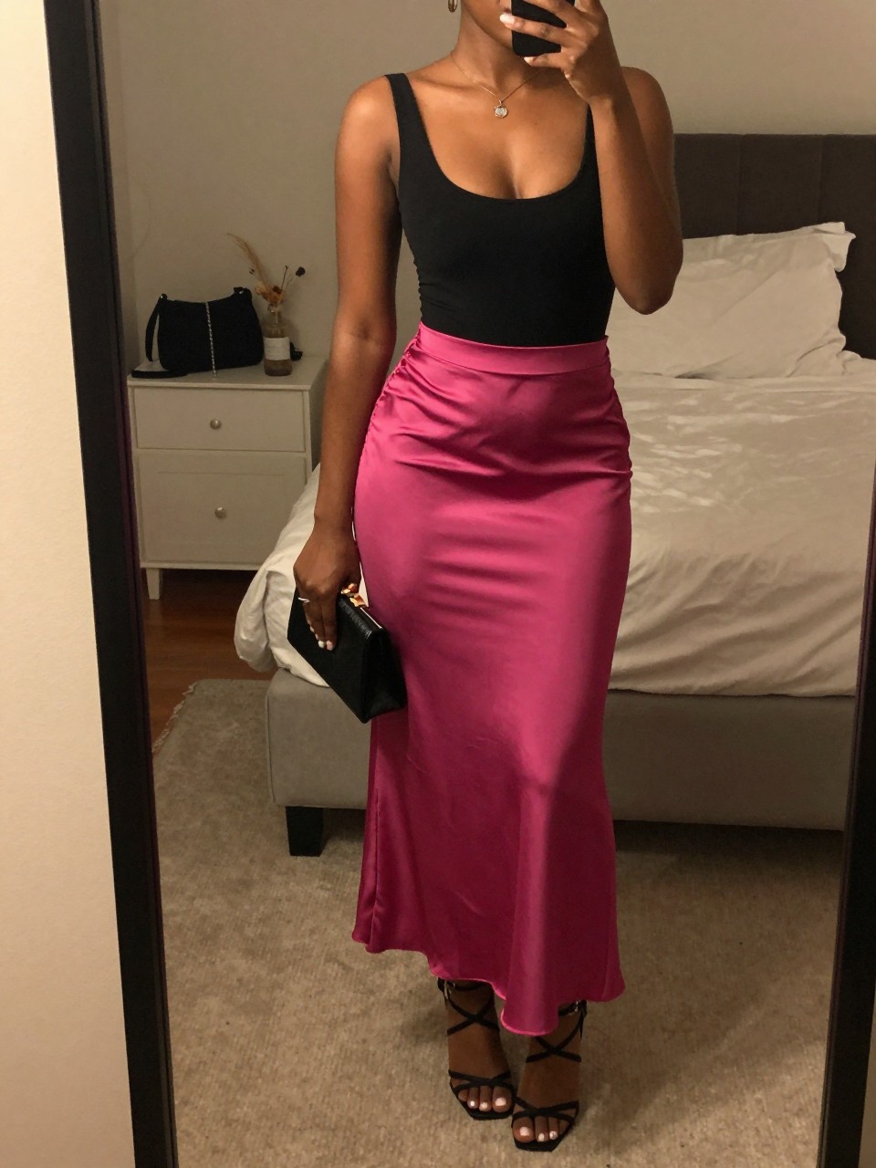 mirror selfie of a black woman wearing a black scoop-neck bodysuit tucked into a magenta pink satin maxi skirt with black strappy heeled sandals and a small black clutch, standing in an elegant evening pose, cropped at NECK so face doesn't show, in a feminine-themed bedroom, warm moody lighting, iPhone photo quality.