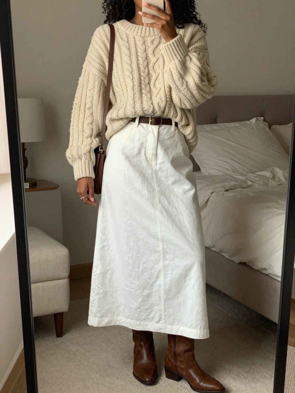 mirror selfie of a black woman wearing an oversized cream chunky cable-knit sweater loosely tucked into a white linen maxi skirt with a brown leather belt and matching brown ankle boots, holding a brown crossbody bag, standing in a cozy elegant pose, cropped at NECK so face doesn't show, in a feminine-themed bedroom, warm moody lighting, iPhone photo quality.