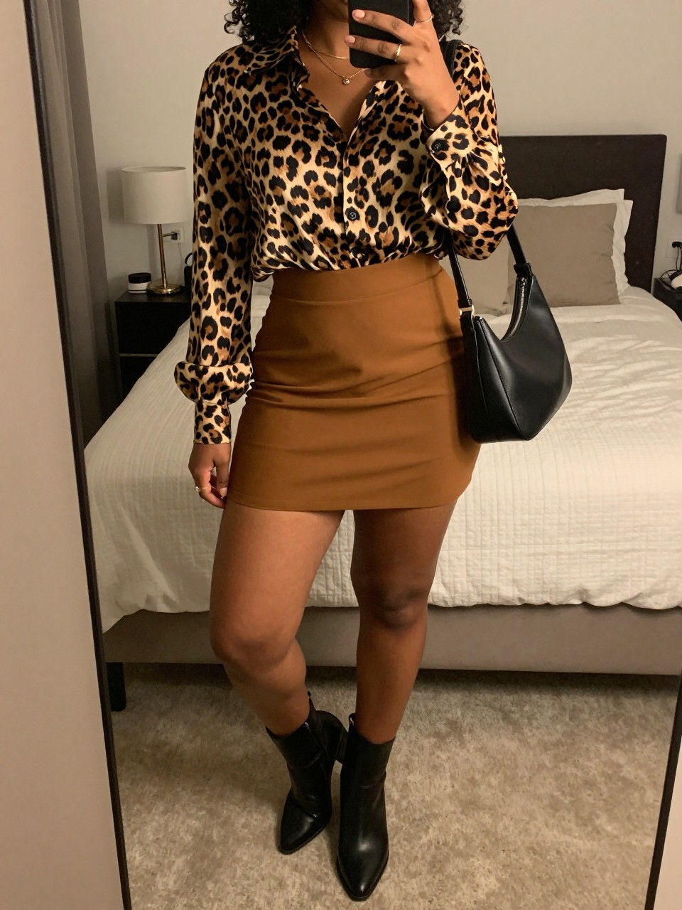 iPhone mirror selfie of a curvy black woman wearing a leopard print silk blouse tucked into a caramel brown A-line mini skirt with black leather ankle boots and a black structured shoulder bag, standing with her weight on one leg, cropped at NECK so face doesn't show, in a feminine-themed bedroom, warm moody lighting, iPhone photo quality.