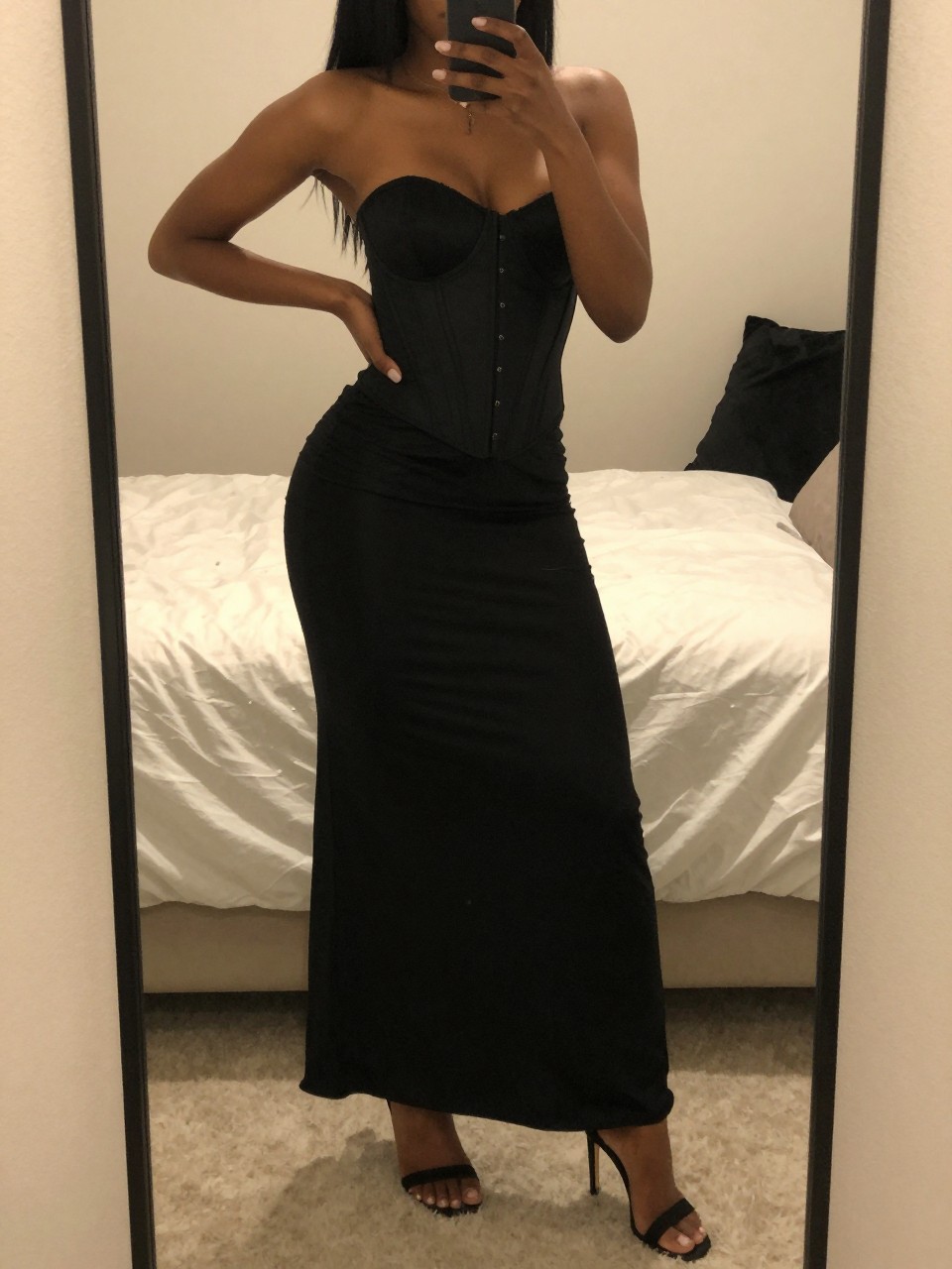 iPhone mirror selfie of a black woman in a black maxi skirt and corset top with heeled sandals, standing with hand on hip, cropped at NECK so face doesn't show, in a feminine-themed bedroom, warm moody lighting, iPhone photo quality.