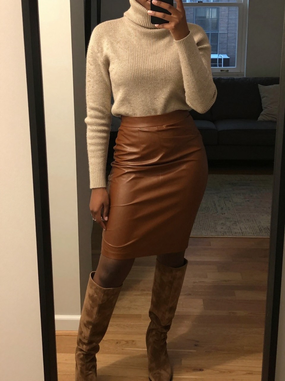 mirror selfie of a black woman wearing a chunky oatmeal turtleneck tucked into a brown leather pencil skirt with knee-high suede boots, standing with her weight on one leg and arms relaxed at her sides, cropped at NECK so face doesn't show, in a city apartment, warm moody lighting, iPhone photo quality.