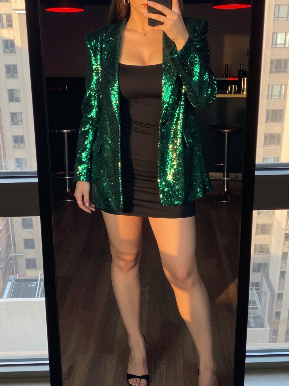 mirror selfie of a curvy woman in a green sequin blazer over a black mini dress with heels, standing in a club or bar, cropped at NECK so face doesn't show, in a city apartment, evening golden hour, iPhone photo quality.
