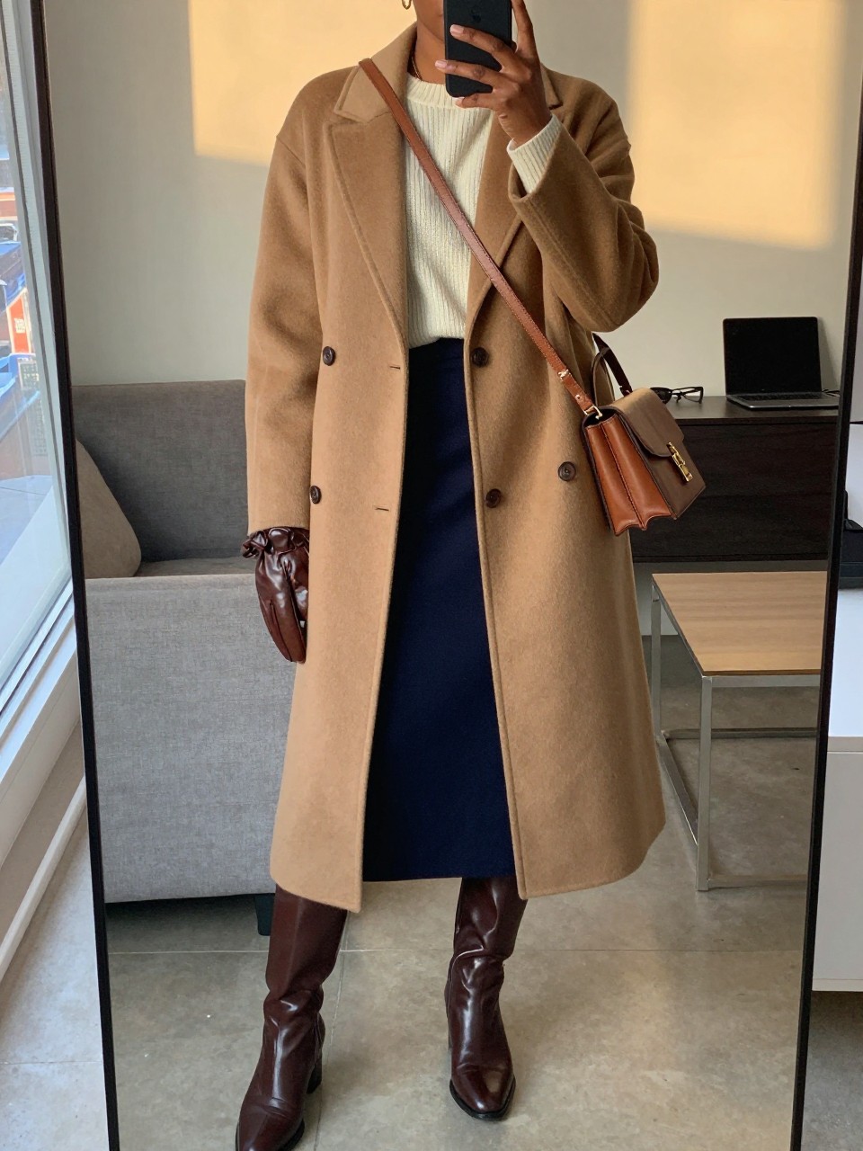 mirror selfie of a black woman wearing a long camel wool coat over a cream sweater tucked into a navy blue midi skirt with brown leather knee-high boots and a matching brown crossbody bag, holding leather gloves, standing in an elegant winter pose, cropped at NECK so face doesn't show, in a city apartment, evening golden hour, iPhone photo quality.