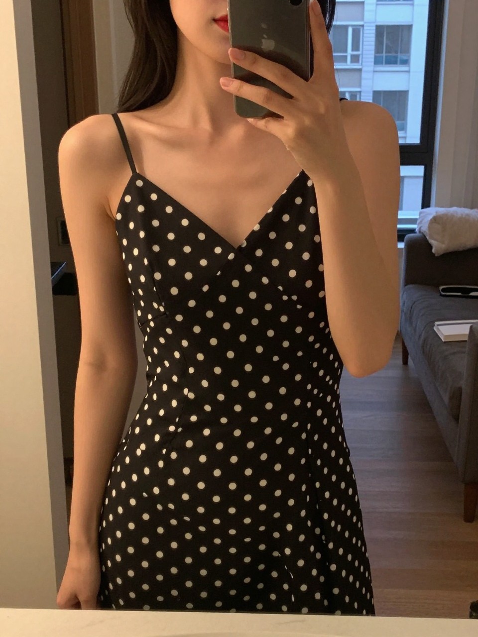 iPhone mirror selfie of a woman in a polka dot dress, applying red lipstick, cropped at NECK so face doesn't show, in a city apartment, warm moody lighting, iPhone photo quality.