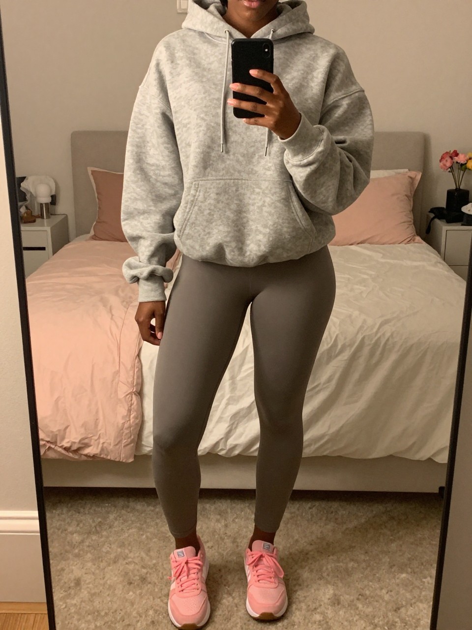 iPhone mirror selfie of a black woman in an athleisure set with an oversized hoodie and pink sneakers, holding phone at chest, cropped at NECK so face doesn't show, in a feminine-themed bedroom, warm moody lighting, iPhone photo quality.